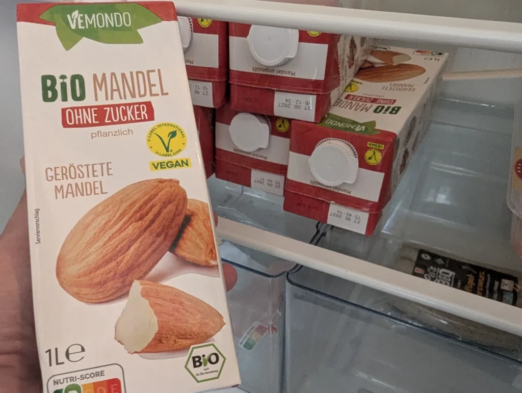Hand hält einen Karton ungesüßte Mandelmilch vor einem Kühlschrank voller Kartons